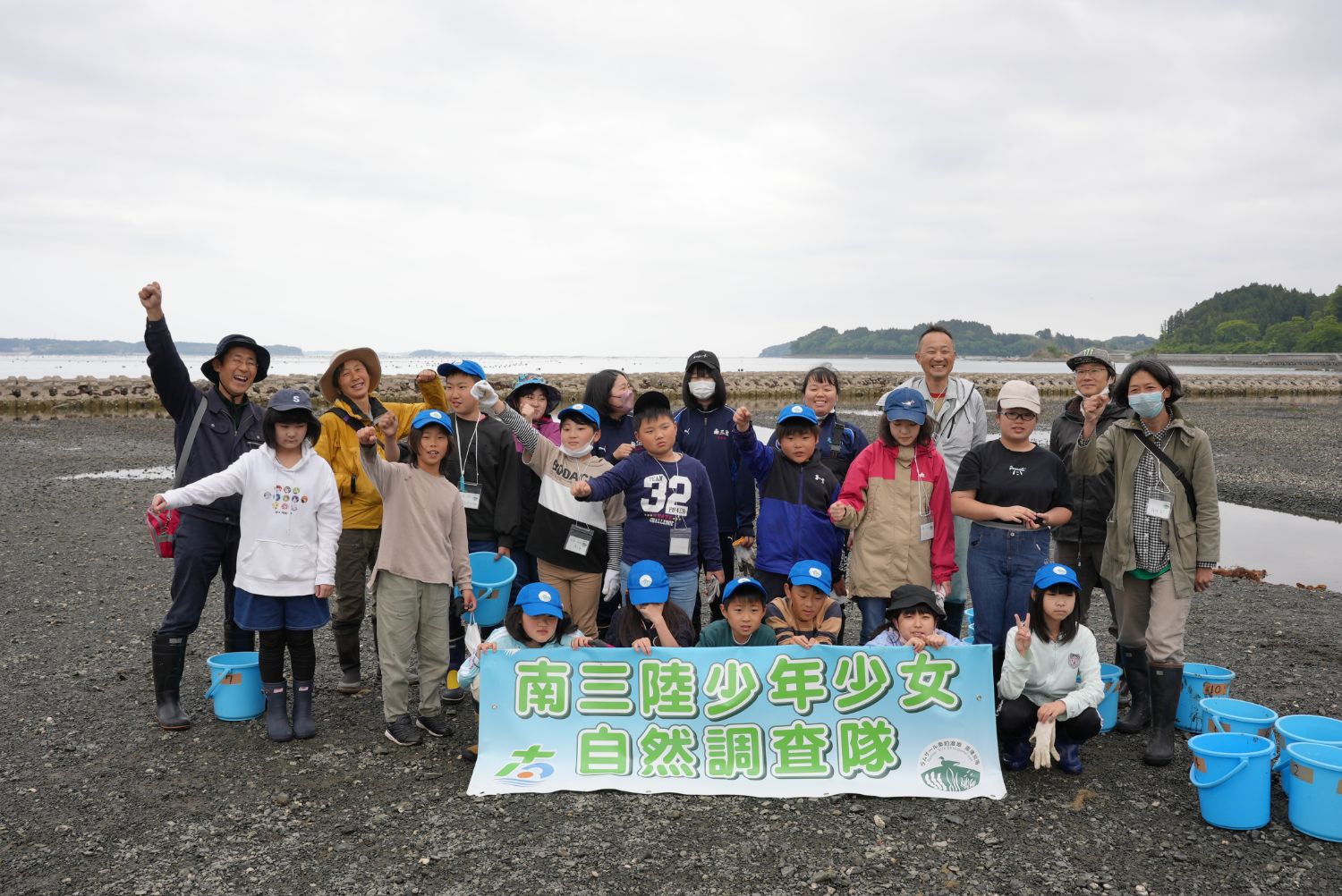 海辺で「南三陸少年少女自然調査隊」の横断幕を掲げ、青い帽子をかぶった子どもたち、大人のスタッフたちが笑顔で並んだ集合写真