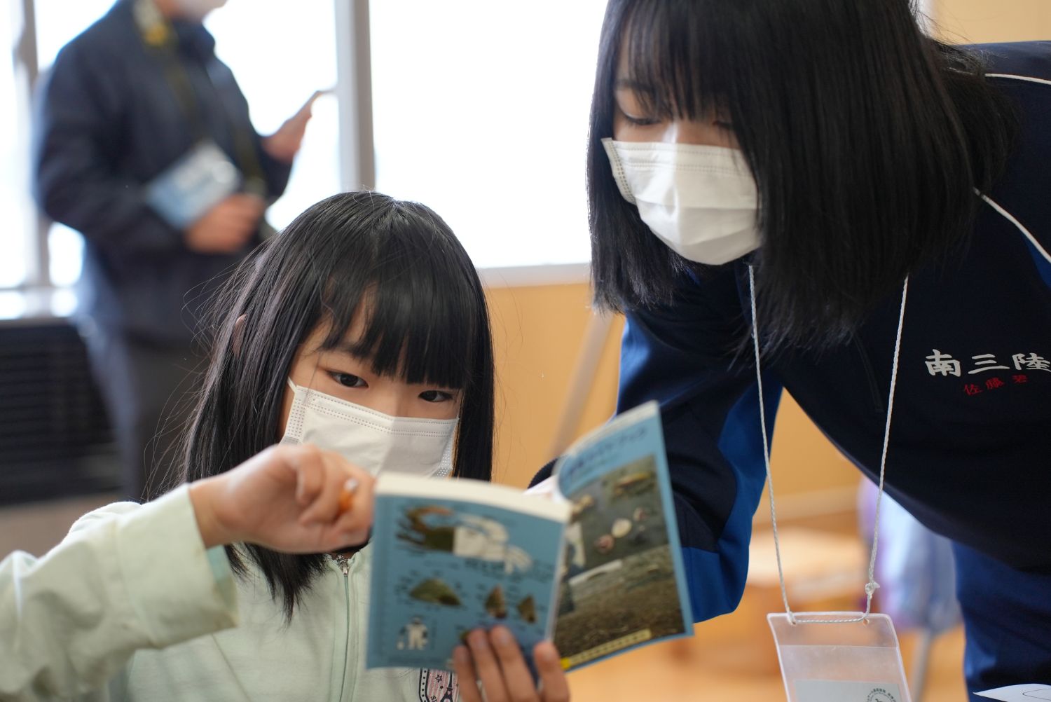 室内で、児童が青い表紙の図鑑を指さしながら読み、横からOGが覗き込んでいる写真