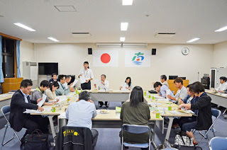 日の丸と南三陸町町旗が並んで掲げられた会議室でコの字型に並べた長机を囲み、関係者の方々が資料を見ながら話し合っている様子の写真