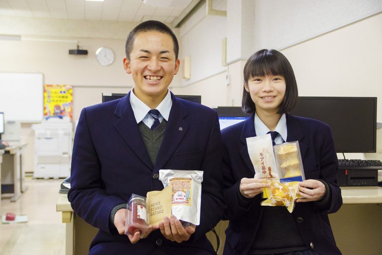 パソコンが設置された教室内で、最中などの沢山の商品を両手で持つ、紺色の制服姿の男子生徒と女子生徒が並んでいる写真