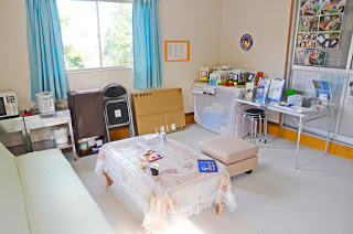 窓から光が差し込む明るい室内で、中央にレースのクロスがかけられたローテーブルと緑色のソファがあり、壁際には電気ポットや折りたたみ椅子、観光パンフレットや食品の展示がある室内の写真