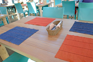 木製のテーブルにオレンジと青のランチョンマットが4枚敷かれ、中央にはカトラリーを入れたカゴが置かれている写真