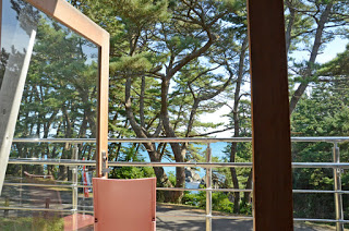 開け放たれたガラス戸の向こうに松林が広がり、その奥に青い海が見えるレストランのテラスからの風景の写真