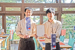 レストランの店内で、白いシャツとストライプのエプロンを着た男性スタッフ2人が「南三陸」と書かれた旗と表彰状を持っている写真