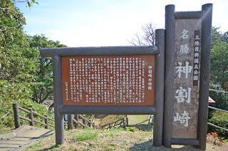 三陸復興国立公園内にある「神割崎(かみわりざき)」という景勝地を紹介するために設置された茶色い案内板と、縦に「名勝 神割崎」と書かれた大きな木製の標識が写っており、背景には階段や木々、海へと続く下り坂の風景が広がっている写真
