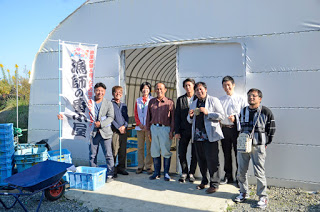 白いビニールハウスのような建物の前でスーツや作業着を着た大人の男性たちが横に並んで立っており、左側には青いプラスチックのコンテナや一輪車が置かれていて、建物の入り口の横には「漁師の番小屋」と書かれた旗が立っている写真