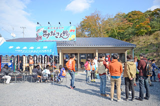 建物「みなと食堂」の前にたくさんの人が集まっており、屋外に設けられたテーブル席では人々が食事を楽しんでいる様子の写真