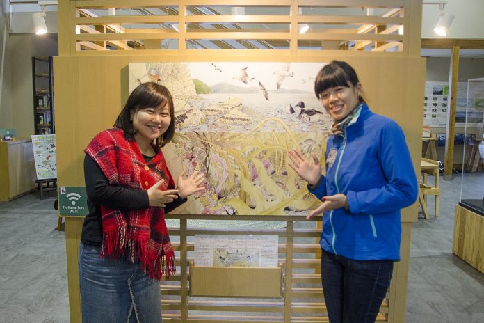 リポーターの女性と青いジャンパーを着た畠山さんが、鳥や自然を描いた展示パネルの前で笑顔で手を広げて紹介している写真