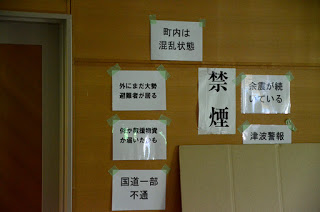 「町内は混乱状態」「禁煙」「外にまた大勢避難者が居る」「救援物資が届いたかも」「国道一部不通」「余震が続いている」「津波警報」のメモが壁に貼られている写真