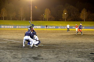 ナイターの野球場でピッチャーが球を投げ、バッターが構えている様子を写した写真