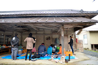 古い木造の建物の広い軒下に青いビニールシートを敷き、そこに座った参加者たちが作業をしている様子を周りに立った数名が見学している様子の写真