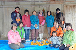 皮をむいてロープに結んだ柿がたくさん置かれたブルーシートの上で、作業を終えた講師の女性たちと参加者の方々が笑顔で記念撮影している写真