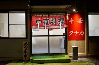 「中華料理」と書かれた赤い暖簾が掲げられ、右側には「中華 タナカ」と書かれた赤い看板、左側には「営業中」と書かれた木製の立て看板が置かれている中華料理店の入口の写真