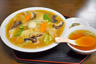 トレーの上に、キノコ・ニンジン・タケノコなどの野菜がたっぷり入った器と、スプーン付きの小さなスープの器が並べられている五目焼きそばの写真