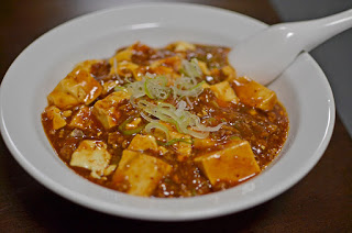 白い陶器のスプーンが添えられた器に、豆腐とひき肉が入った麻婆豆腐が盛られ、刻んだ青ねぎが上に散らされている写真