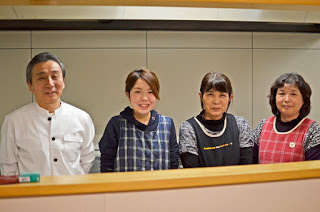 店主は黒いボタン付きの白いシャツを着ていて、他の3人はそれぞれ青・黒・赤のチェック柄のエプロンを着用している、スタッフ4人の人物が並んでいる様子をカウンター越しに撮影した写真