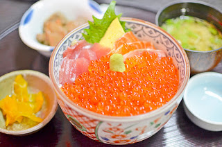 白いご飯の上に鮮やかなオレンジ色のイクラ(鮭の卵)がたっぷりと盛られ上には少量のわさびが載っていて、いくら丼の横には味噌汁や漬物、煮物などが小鉢に入って並べられた料理の写真