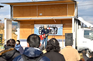 屋外のイベント会場で、「宮城県南三陸町復興市」と書かれた横断幕が飾られたトラックの荷台に設けられたステージ上で、エレキギターを演奏する人物と、それを見ている観客の後ろ姿を写したライブ演奏の写真