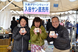 イベント会場の「入谷グリーンウェーブ 入谷サン直売所」と書かれた看板が掲げられているテントの下で、3人の男女が小さな箱の商品を手に持ち、カメラに向かって見せている写真