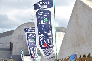 白いテントが張られた屋外の会場で「がんばろう!南三陸 復興市」と書かれたのぼり旗が風になびいている写真