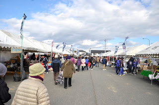 白い雲がぶ青空の下、左右にずらりと並んだ白いテントの間の通路を多くの来場者が歩き、テントで品物を見たりしているにぎやかな復興市の様子の写真