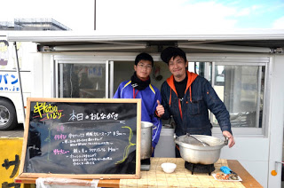 二人の男性の前のテーブルに、調理器具と大きな鍋が並び、本日のおしながきと書かれた黒板が置かれている屋台の写真