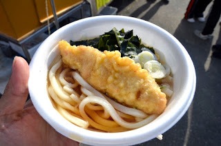 屋外の風景を背景に、手に持たれた白い器の中に天ぷら、わかめ、刻みネギがのったうどんが盛られている、温かい料理の写真