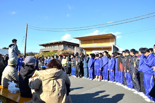 青空の下、青いジャージ姿の生徒たちが並んで立ち、向かい合って設置された台の上に立つ男性の方を見ている様子を、手前で座って見守る人々とともに写した写真