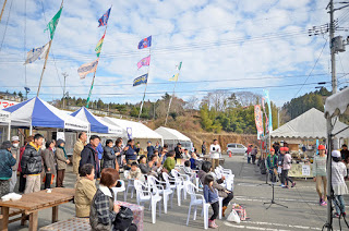 青空の下、屋外のイベント会場でテントや旗が並び、多くの人々が椅子に座ってステージを見守っているにぎやかなイベントの写真