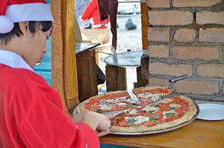 レンガ壁の前で、赤いサンタクロースの衣装と帽子を身につけた男性が、木の台の上で大きなピザを切っている様子の写真