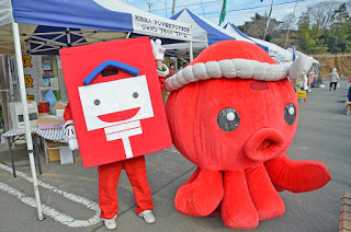 屋外のイベント会場で、白い顔の下に郵便マークが描かれた赤いポストのキャラクターと、白いハチマキを巻いた大きな赤いタコのキャラクターが並んで立っている写真