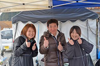 屋外のテントの下で、茶色いジャケットの男性の左右に女性が2人立ち、そろって親指を立てるサムズアップのポーズをして並んでいる写真