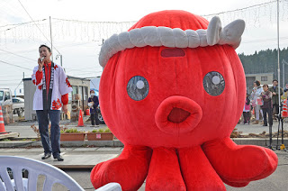 屋外のイベント会場で、白いハチマキを巻いた大きな赤いタコのマスコットの後ろのステージで白い法被を着た男性がマイクを持ち歌を歌っているイベントの様子の写真