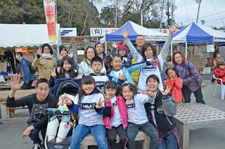 屋外に設置されたテントや屋台の前で、ベンチに座る子どもや大人の後ろに人々が集まり、カメラに向かって手を振ったりピースサインをしている集合写真