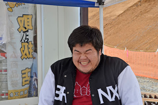 屋外のテントの下で、赤いシャツの上に黒と白のジャケットを着た男性が満面の笑顔で写っている写真