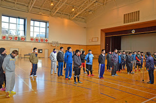 体育館の中でマイクスタンドの前に立つ人の話を、ジャージ姿の多くの人々が整然と並んで立ち聞いている様子の写真