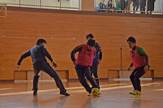 2階の観客席から人が見ている屋内コートで、ピンクのビブスを着た2人と黒いジャケットを着た2人の計4人が、黄色と黒の模様のサッカーボールを使ってプレーしている様子の写真