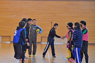 体育館の中で、中央にいる男性と右側の先頭に並んでいる人が握手を交わし、周囲にスポーツウェアを着た他の人々が立って見守っている様子が写されている写真