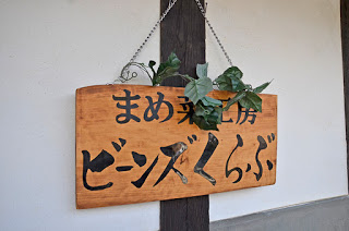 「まめ菜工房ビーンズくらぶ」と書かれ、緑色のツタが飾られた木製の看板が、白い壁の木の柱にチェーンで掛けられた写真