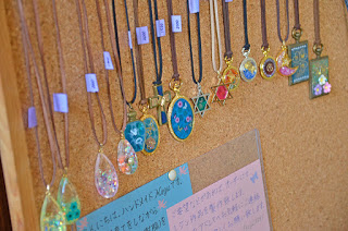 ハンドメイド作家の手書きのメッセージが書かれた紙が貼られたコルクボードに、各ネックレスにラベルが付けられた、涙型や円形など様々なデザインのペンダントが付いたネックレスが並んでいる様子の写真