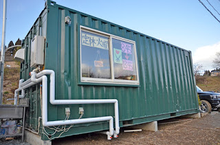 緑色の貨物用コンテナを改装して建てられた小さな建物で、側面の窓にはカラフルな紙に手書きされた営業情報やイラストが貼られている美容室「Rotti(ロッティー)」の外観写真