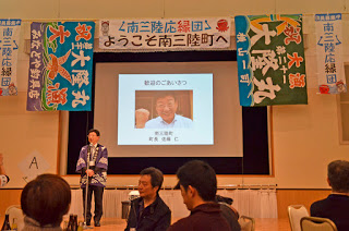 出席者たちがテーブル席に着席している会場で、「南三陸応縁団ようこそ南三陸町へ」と書かれた横断幕や、2枚の大漁旗に囲まれた大きなスクリーンに男性の顔写真が映し出され、その前に立った紫色の法被を着た男性が、マイクで話をしている写真