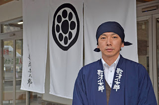 紺色の布を頭に巻き、同じ色の「歌津小太郎」と書かれた法被を身につけた男性が、丸の中に点を配した紋が描かれた白いのれんの前で、正面を向いて立っている写真