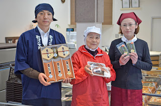 ショーケースの前で、左の紺色の法被を着て同じ色の布を頭に巻いた男性が箱入りの商品、中央の赤い作業着と白い帽子をかぶった女性が食品トレーに入った商品、右のえんじ色のエプロンと三角巾を着用した女性が透明パック入りの商品を持ち、3人並んで笑顔で立っている写真