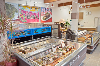 明るく清潔感のある空間に加工食品を並べたショーケースが2つ並び、奥にある水槽の上には「福福丸」と書かれた大漁旗や加工品の写真看板が掲げられている店内の写真