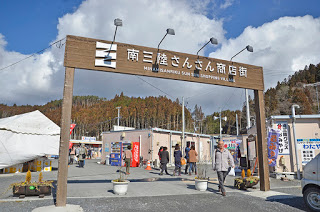 「南三陸さんさん商店街」と白い文字で書かれた、大きな茶色のゲートの中に仮設商店街の建物が建っている写真