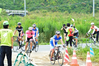 ピンクや青のサイクリングウェアを着た複数の自転車選手が分岐地点で誘導員の指示を受けながら曲がろうとしている様子で、カメラマンが選手を撮影している場面の写真