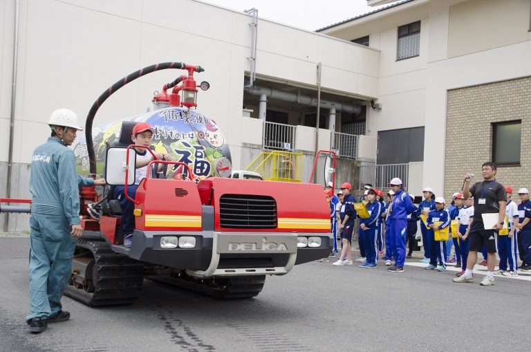 青色のジャージ姿の児童や先生が整列して見守るなか、ヘルメットを被り薄い緑色の作業服を着た男性スタッフの指導のもと、男子児童が後ろにタンクが設置された赤色の作業車の運転席に座っている様子の写真