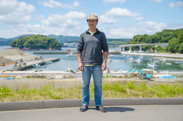 船の停泊する静かな漁港や緑の山々、高架道路が見える風景をバックに立っている佐々木昌則さんの写真