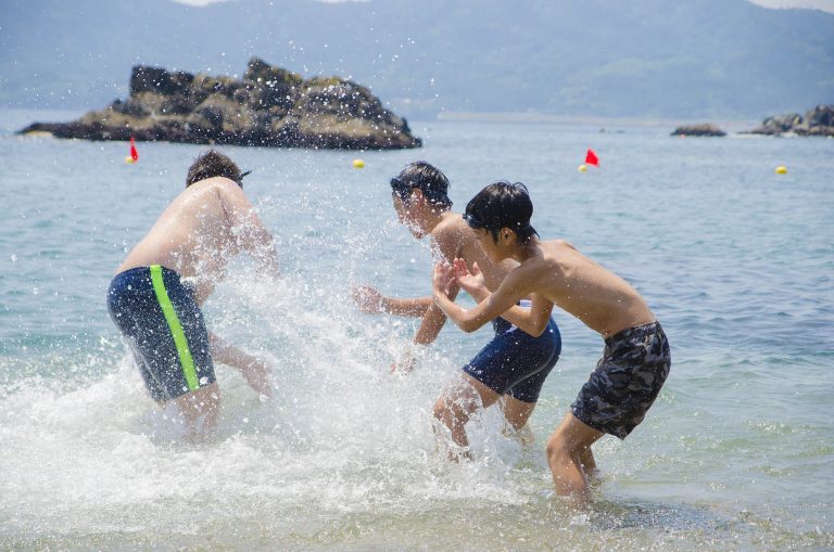 小島があり赤色のフラッグが浮いている海水浴場で、水着を着た3人の男性が水をかけあっている様子の写真